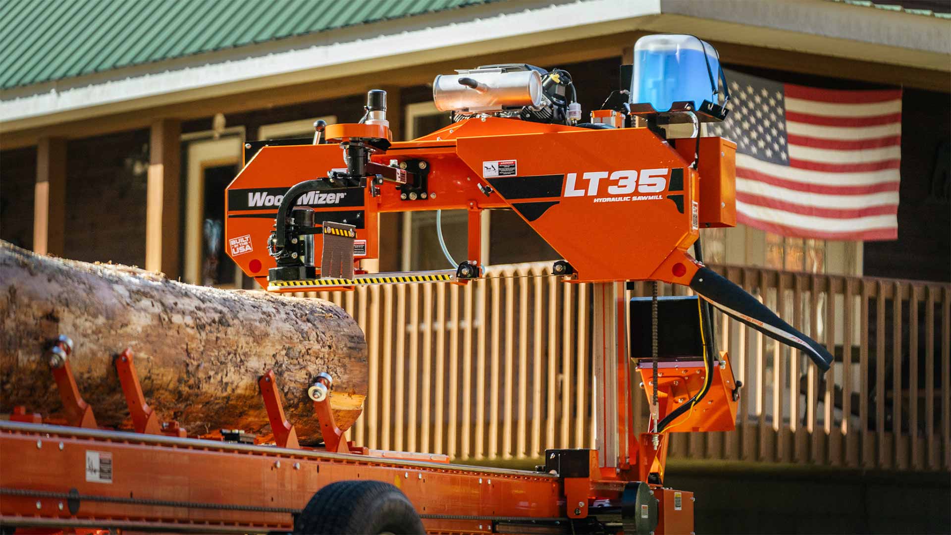 Orange Wood-Mizer LT35 Hydraulic band sawmill with a bark-covered log clamped on its steel bed by hydraulic side supports, the cutting head marked LT35 HYDRAULIC SAWMILL, a blue lubricant tank above it, and a house porch and American flag behind