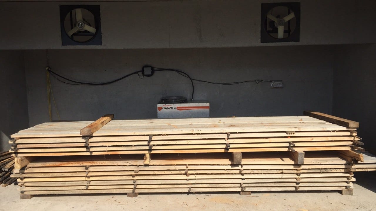 A Wood-Mizer KD250 kiln unit mounted on the concrete back wall of a drying chamber between two wall fans, with a stickered stack of freshly sawn pale boards filling the floor in front of it