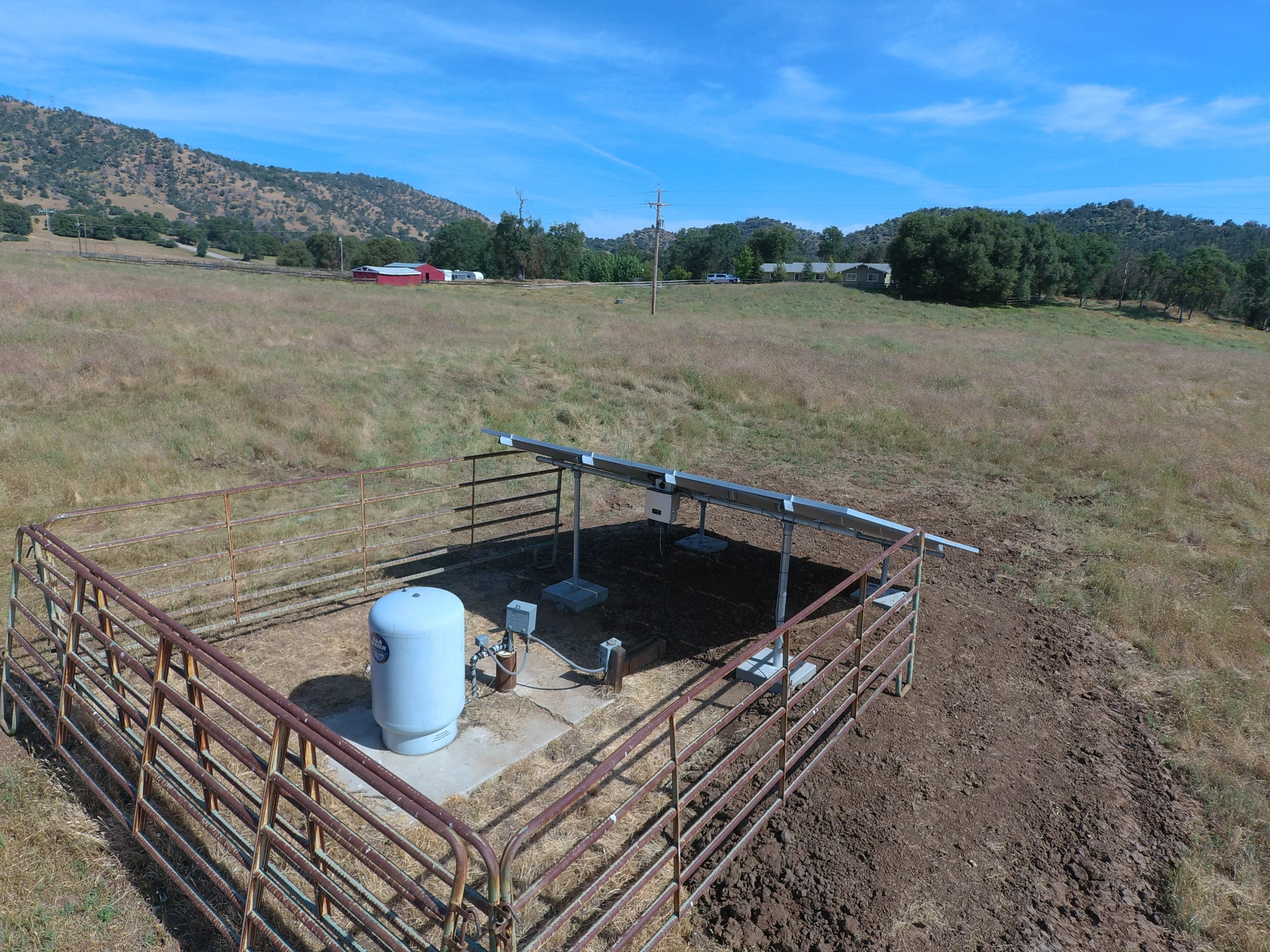 A ground-mounted solar array and a white pressure tank fenced inside corral panels in a dry California pasture, with a distribution power line running along the road behind it