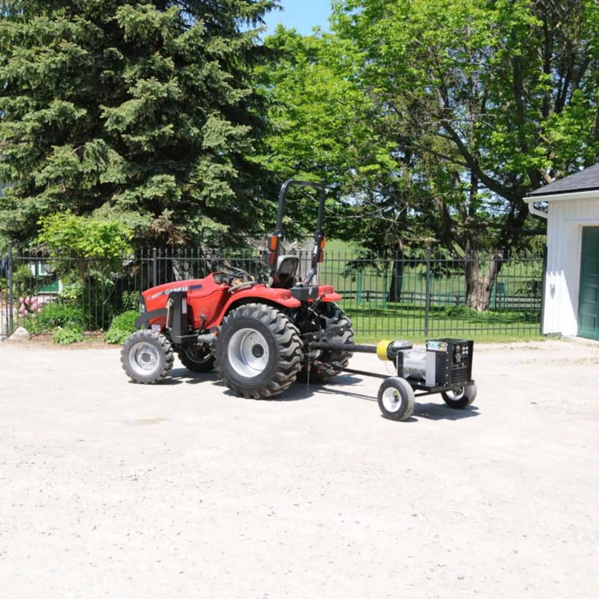 A trailer-mounted Baumalight PTO generator hitched behind a red compact Case IH tractor by its driveshaft, parked on a gravel yard beside a white shed