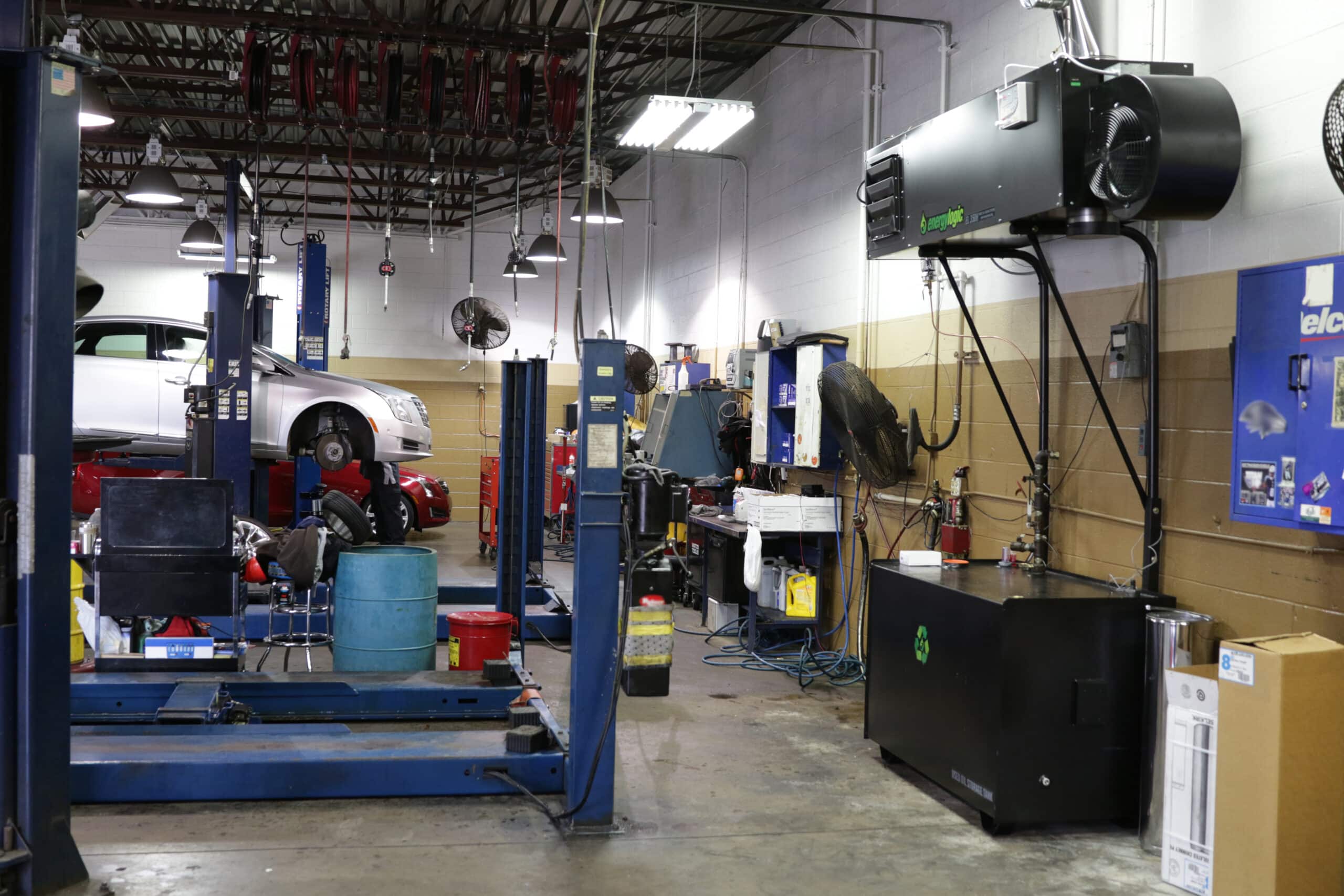 An EnergyLogic waste oil furnace hanging on a steel stand above its black waste-oil storage tank inside a working car dealership service bay, with vehicles on lifts and hose reels overhead