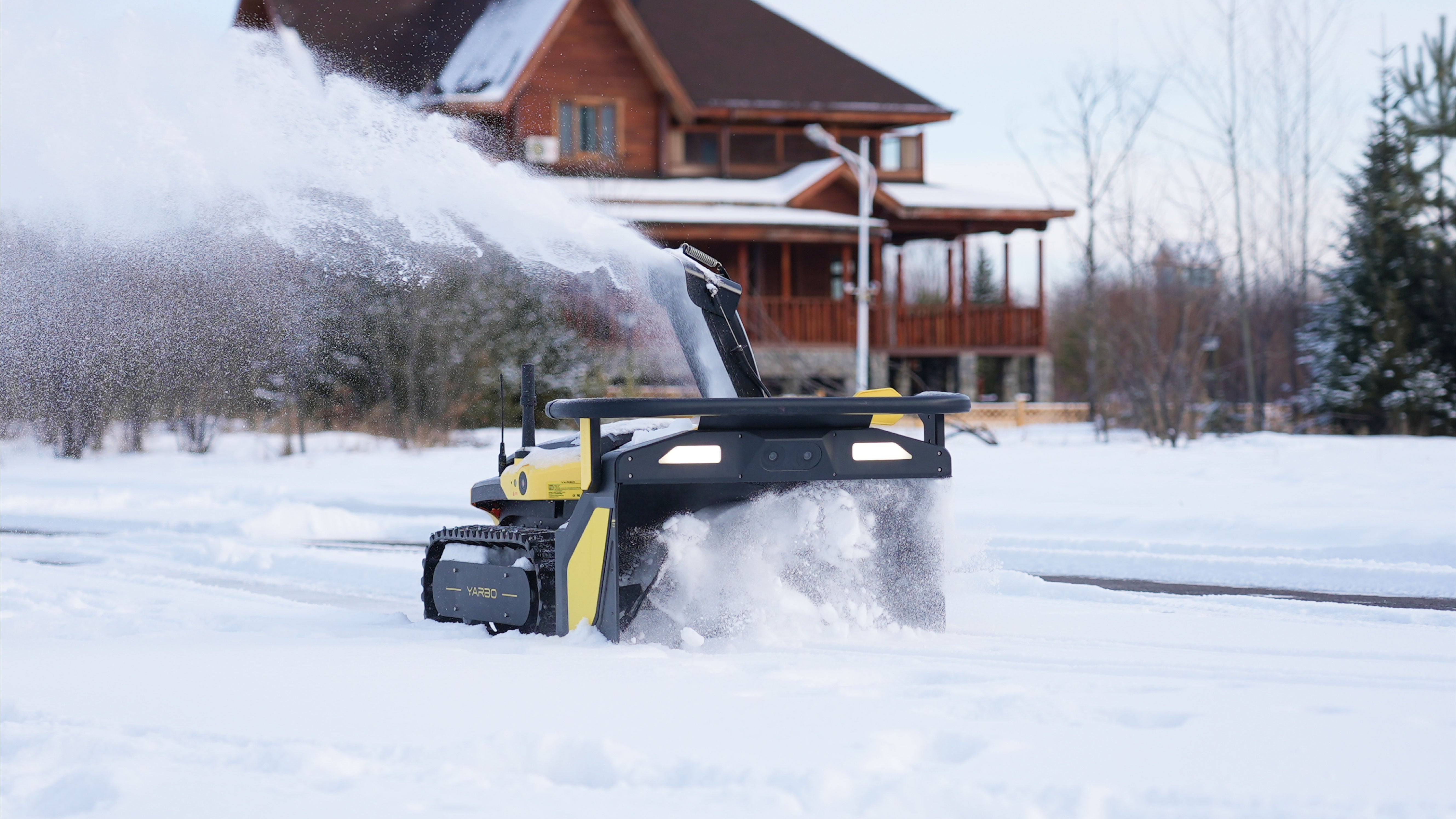 A yellow and black tracked Yarbo robot throwing a long plume of snow across a white driveway in front of a two-story timber house, with snow-covered spruce behind it