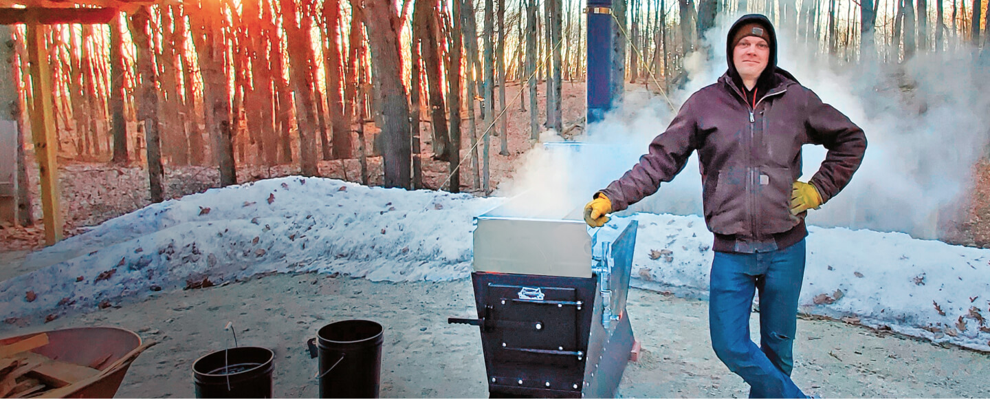 A Smoky Lake Dauntless evaporator boiling in a snowy maple woodlot at dusk, steam pouring off the open pan and out of the stack, with a person in a hooded jacket standing beside it and sap buckets on the ground