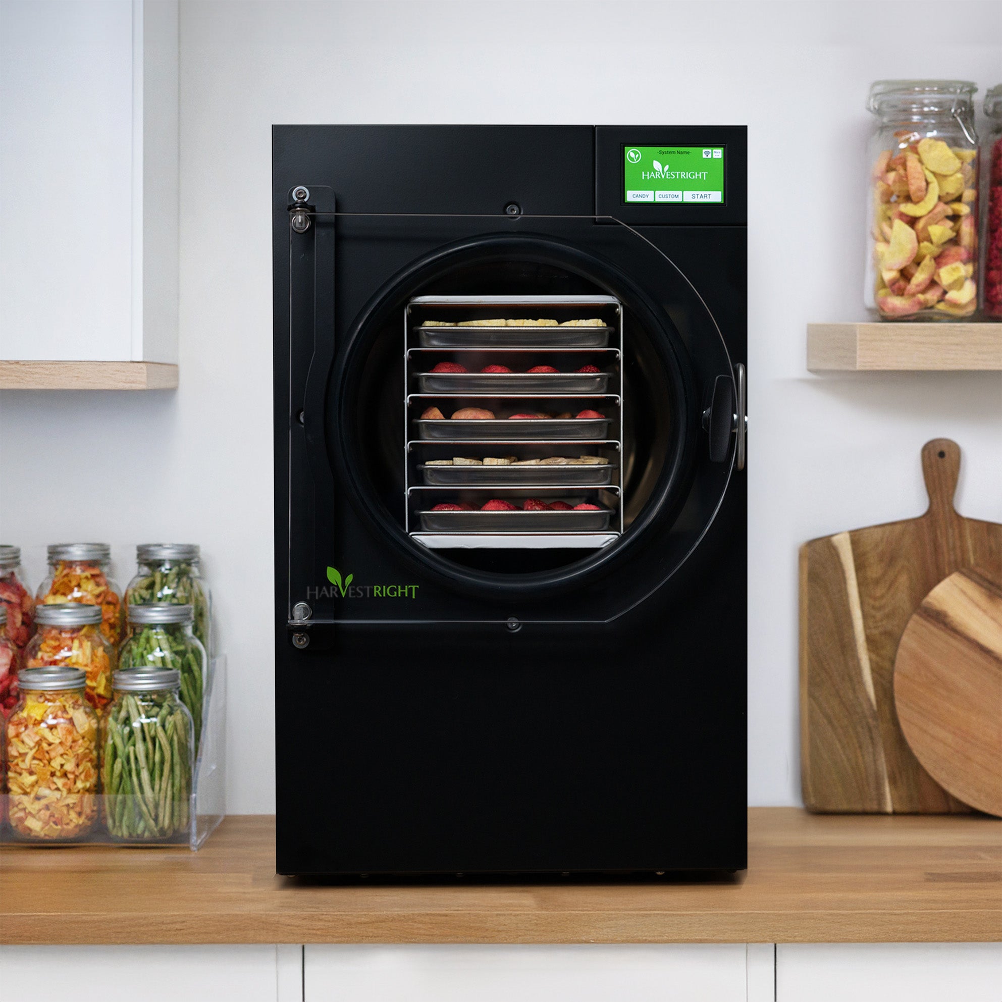 A black Harvest Right Medium home freeze dryer standing on a wooden kitchen counter, five stainless steel trays of strawberries, apples and bananas racked behind its clear door, with jars of finished freeze-dried food on the shelves beside it