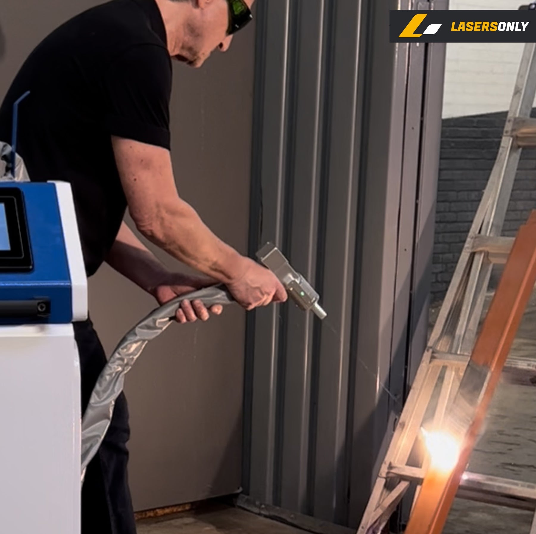An operator in laser safety glasses holding the handheld gun of a CW 2000W fiber laser cleaner, stripping coating off a steel frame inside a shop, with the blue and white machine cabinet and its coiled fiber cable beside him