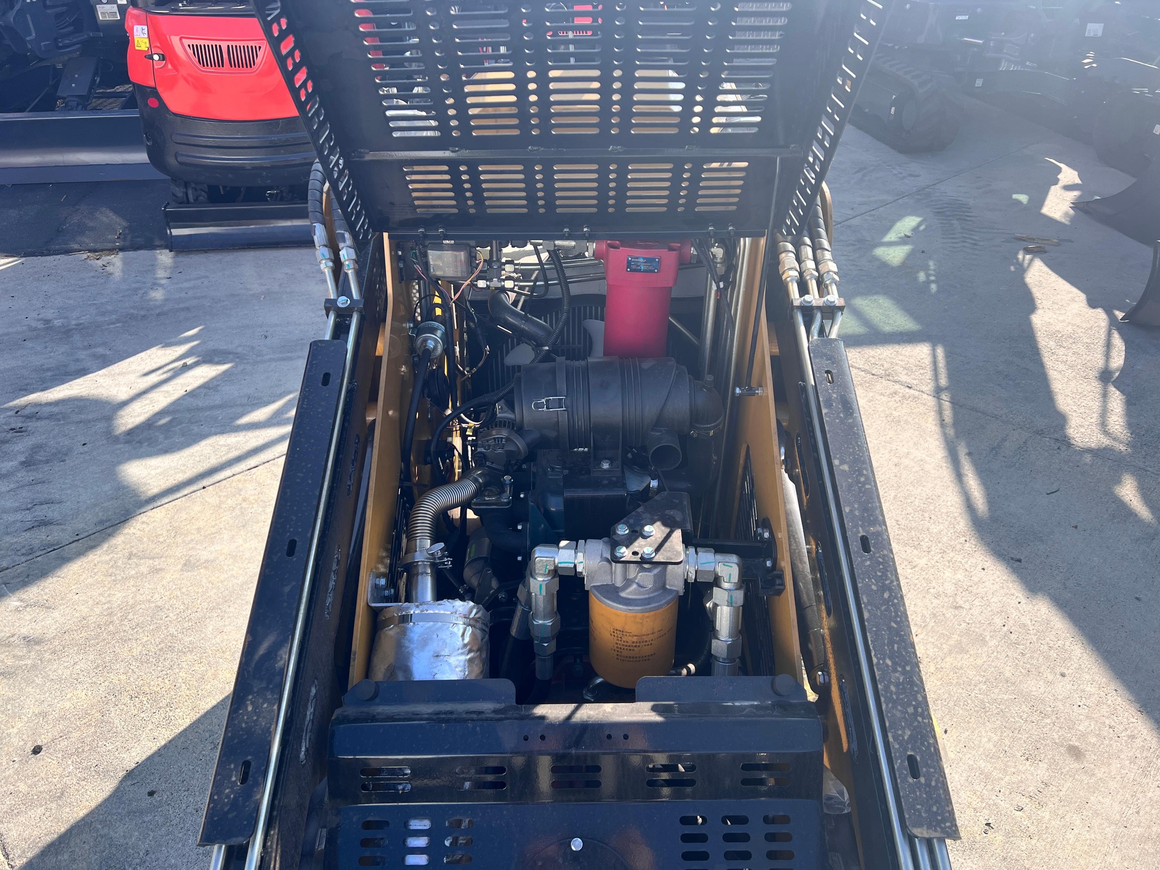 An AGT KKT27 mini skid steer parked on a dealer's concrete yard with its rear hood tilted open, showing the Kubota D1105 diesel, the black air cleaner, a red hydraulic filter and spin-on filters, with a red mini excavator beside it and the photographer's shadow across the slab