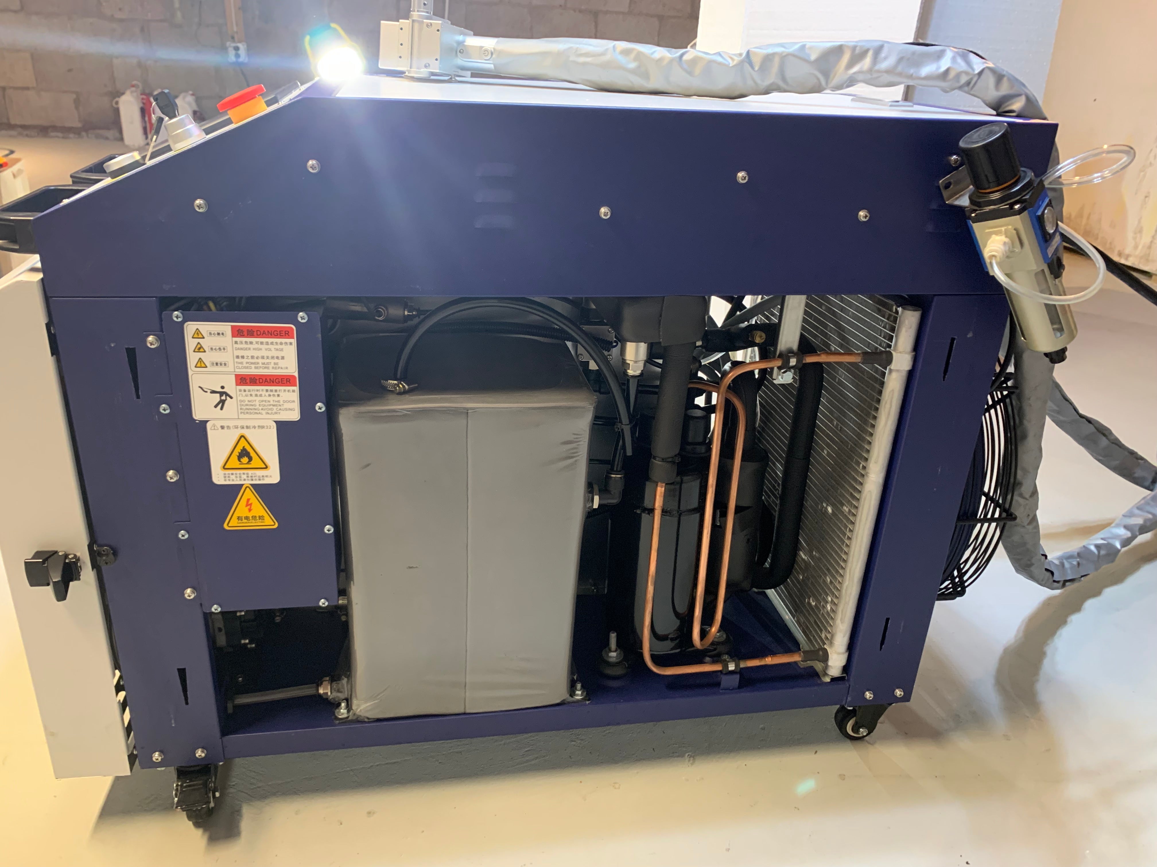 Gabinete azul de un limpiador láser de fibra fotografiado en un taller de paredes de bloque con un panel lateral quitado, a la vista el enfriador de agua con su serpentina, los caños de cobre y el ventilador, el conducto aislado de la fibra enrollado arriba y las etiquetas de peligro láser en el bastidor