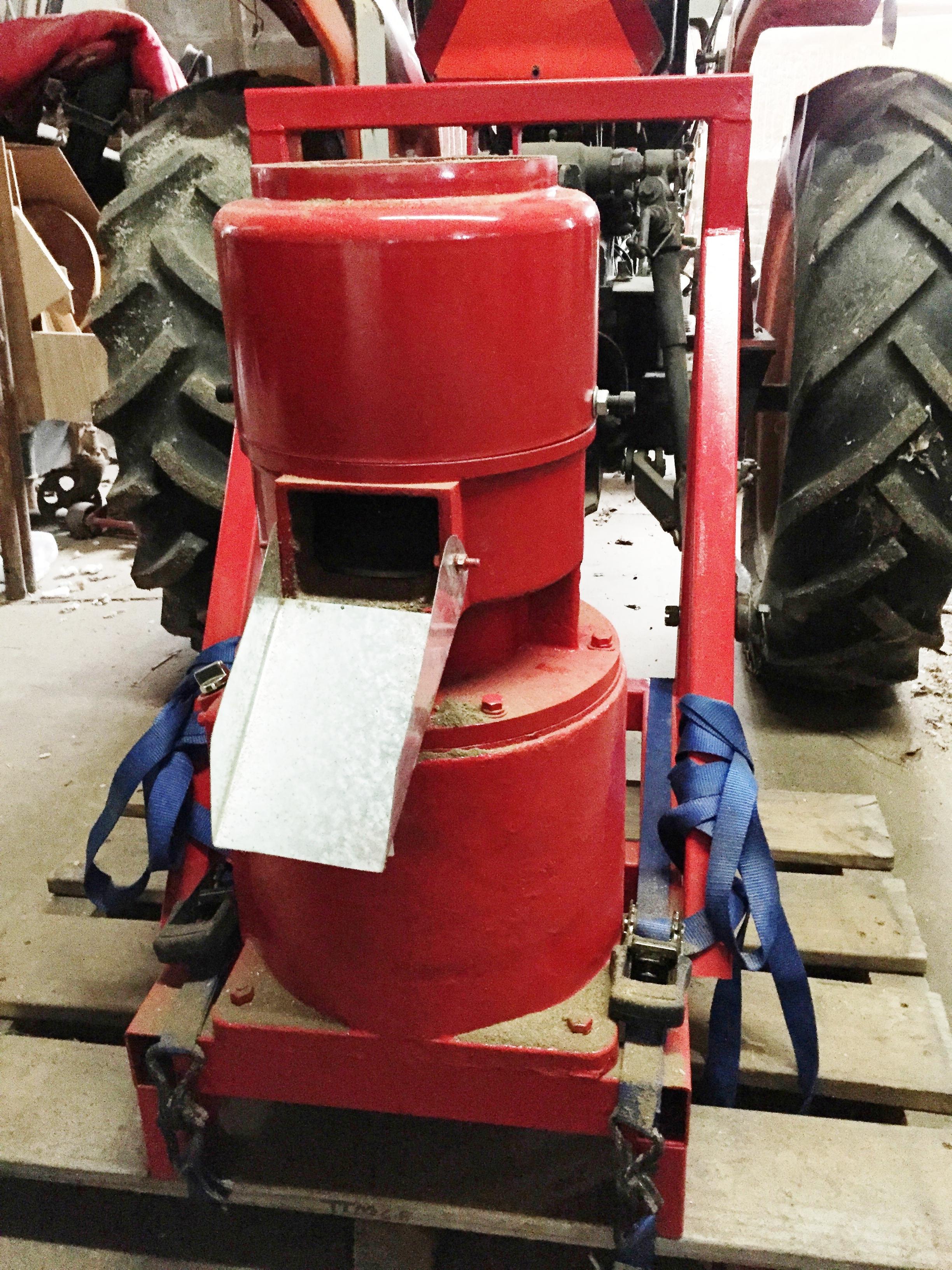 A red PTO-driven flat die pellet mill on its three-point frame, strapped to a wooden pallet and dusted with sawdust, standing on a shop floor between the rear tyres of a tractor with the hitch visible behind it