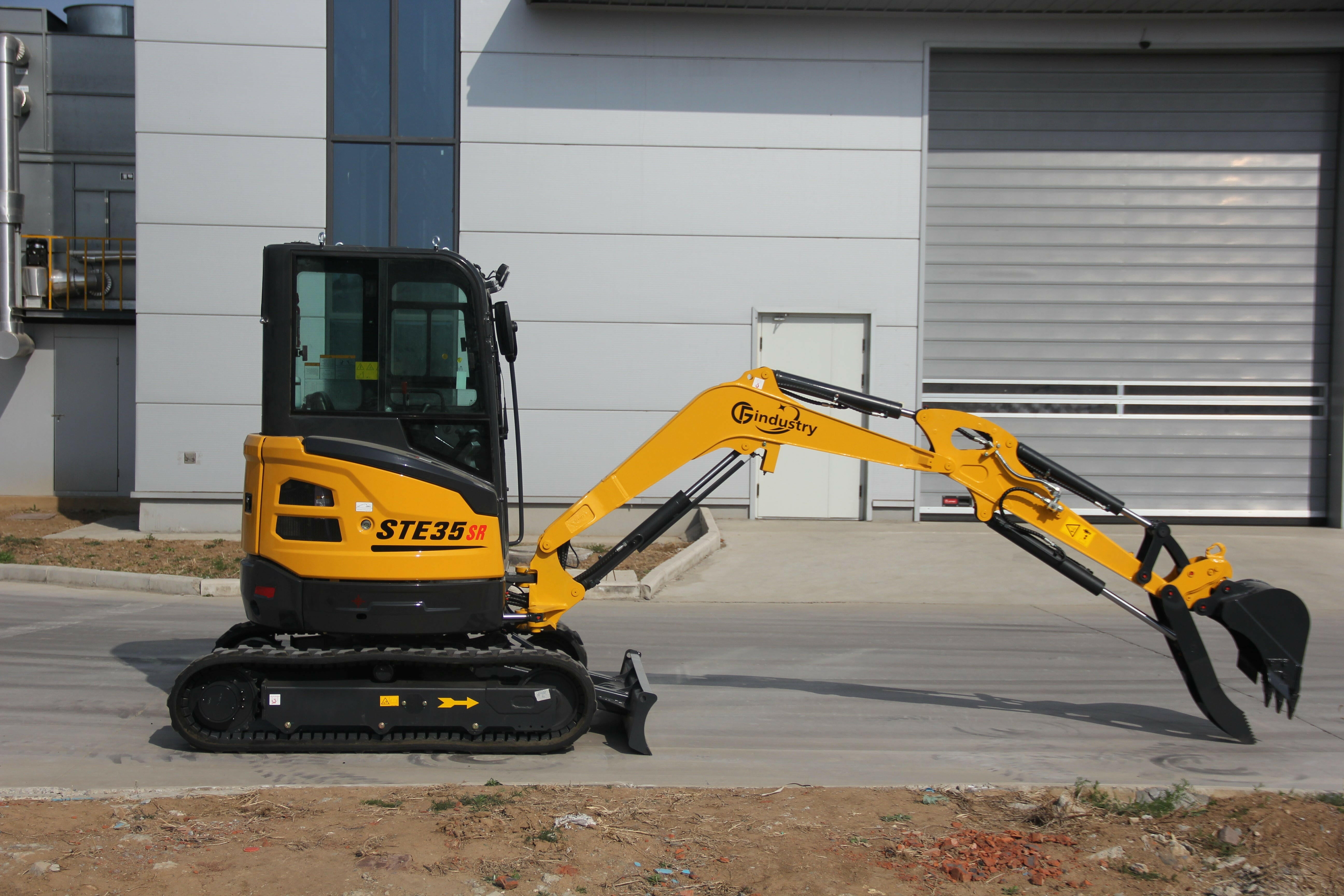 A yellow CFG Industry STE35SR tracked mini excavator standing on concrete outside a grey industrial building, its boom extended to the right with the hydraulic thumb open above the bucket and the dozer blade lowered in front of the tracks